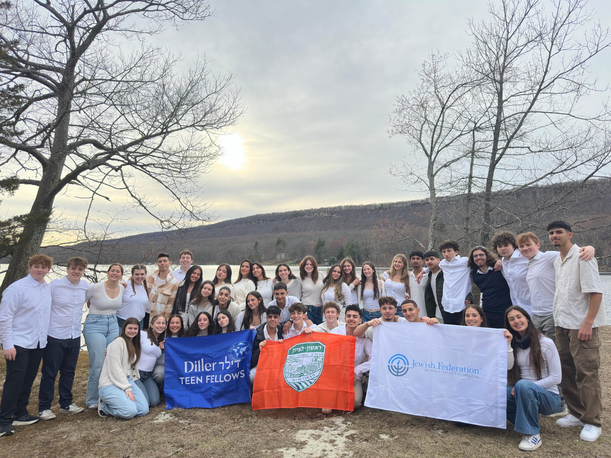 All the teens from the Jewish Community Mifgash standing together in the woods by a lake at sunset, holding Diller Teen Fellows posters and smiling.